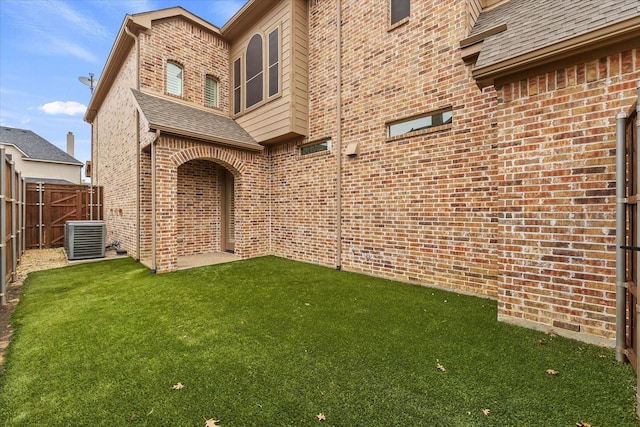 exterior space with a lawn, central air condition unit, brick siding, fence, and a shingled roof