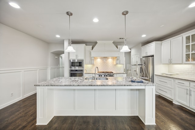 kitchen with a center island with sink, arched walkways, stainless steel appliances, premium range hood, and a sink