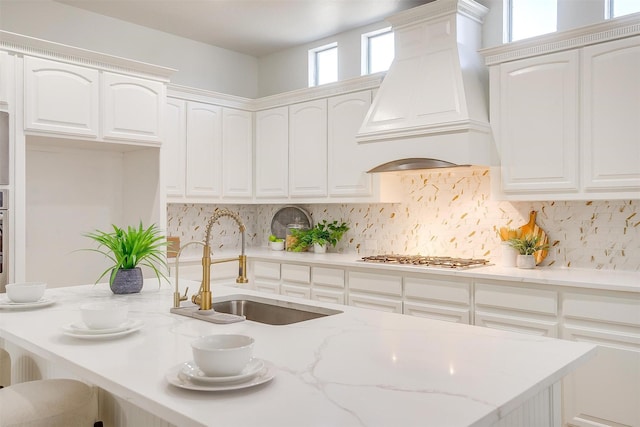 kitchen with stainless steel gas cooktop, tasteful backsplash, custom range hood, white cabinets, and a sink