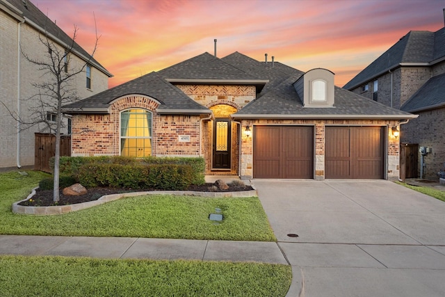 french country inspired facade featuring a garage, brick siding, concrete driveway, stone siding, and roof with shingles