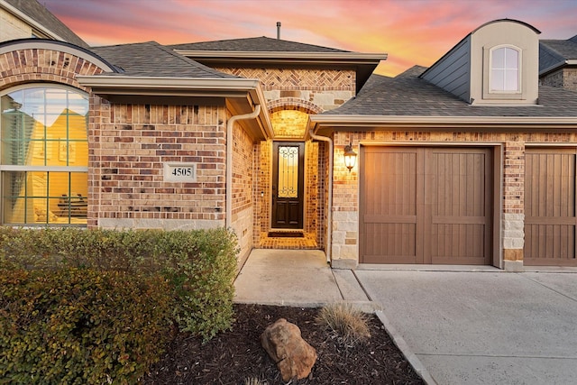 entrance to property with an attached garage, driveway, roof with shingles, and brick siding