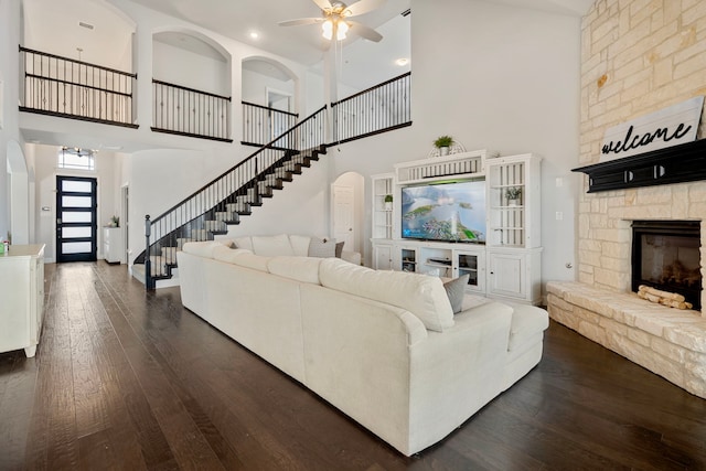 living area with dark wood-style flooring, stairway, a high ceiling, a ceiling fan, and a stone fireplace