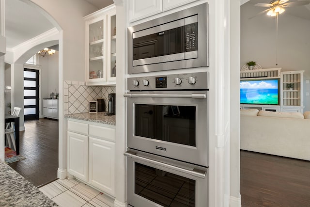 kitchen featuring arched walkways, glass insert cabinets, appliances with stainless steel finishes, light stone countertops, and backsplash