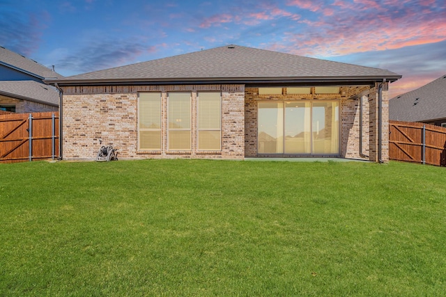 back of house at dusk with brick siding, a lawn, and fence