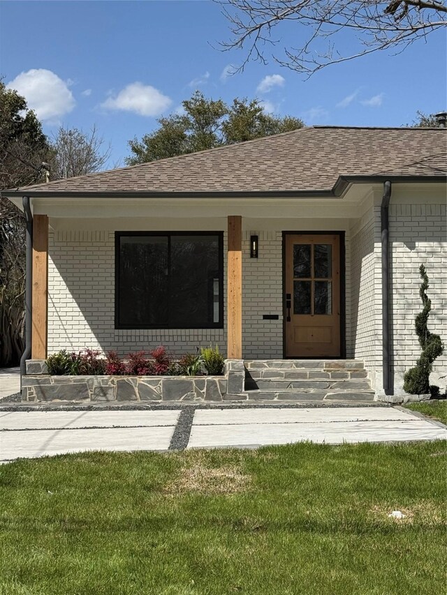 property entrance featuring brick siding, a lawn, a porch, and a shingled roof