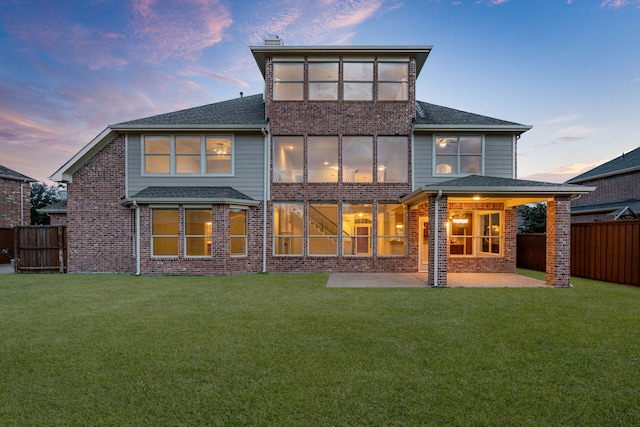 back of property at dusk with fence private yard, a patio area, a lawn, and brick siding