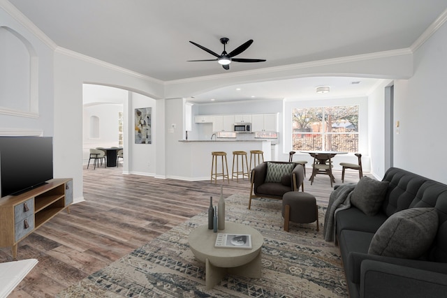 living room with arched walkways, crown molding, ceiling fan, wood finished floors, and baseboards