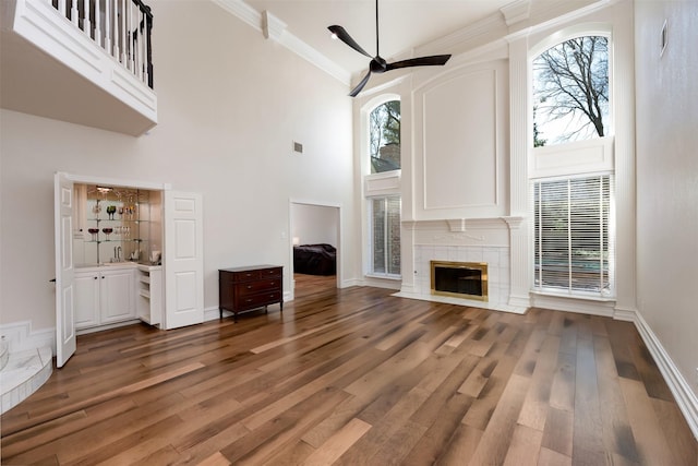 unfurnished living room featuring a tile fireplace, a towering ceiling, ceiling fan, ornamental molding, and wood finished floors