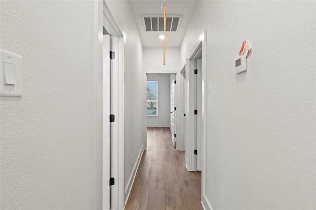 hallway with a textured wall, wood finished floors, visible vents, baseboards, and attic access