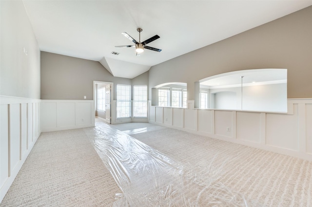 carpeted empty room with lofted ceiling, visible vents, a decorative wall, a ceiling fan, and wainscoting