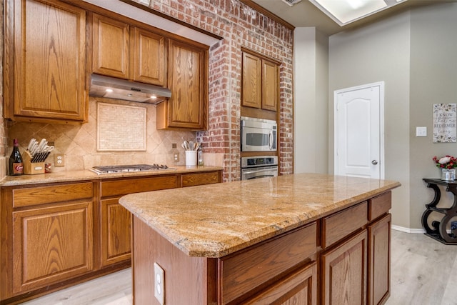 kitchen featuring tasteful backsplash, light wood-style floors, appliances with stainless steel finishes, brown cabinets, and under cabinet range hood