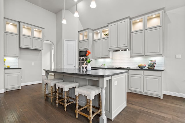 kitchen with dark wood-style floors, arched walkways, a center island with sink, stainless steel appliances, and dark countertops