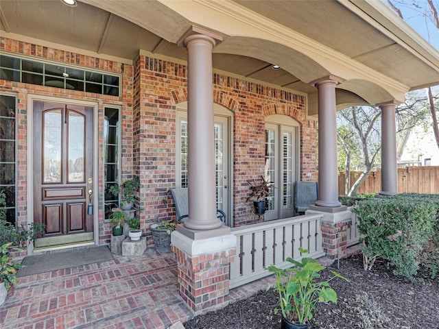 view of exterior entry featuring fence, brick siding, and covered porch