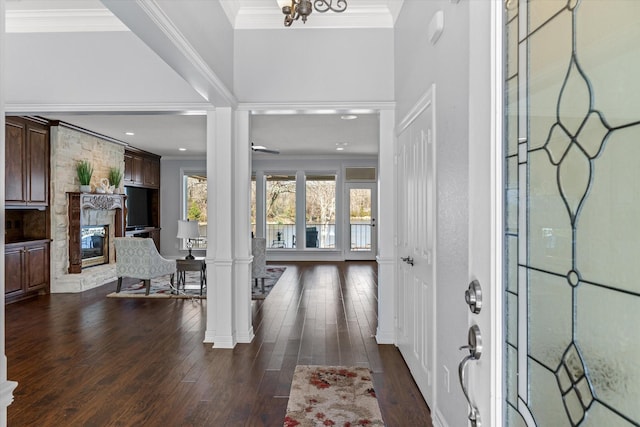 entryway with a stone fireplace, dark wood-style flooring, decorative columns, and crown molding