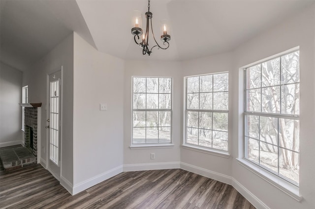 unfurnished dining area with a brick fireplace, baseboards, a notable chandelier, and dark wood-style flooring