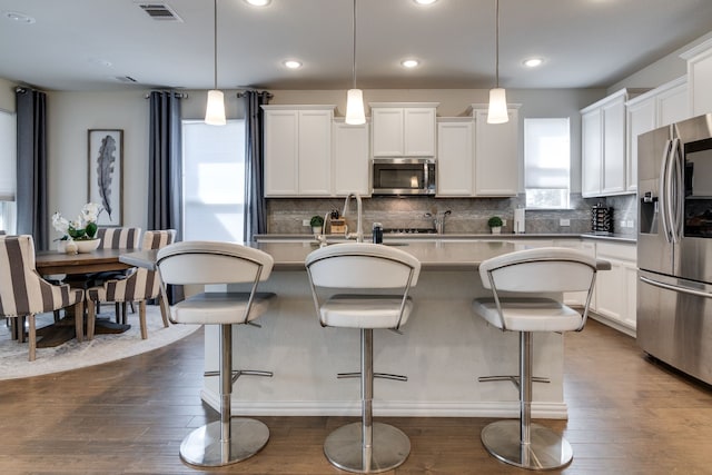 kitchen with dark wood-style flooring, visible vents, appliances with stainless steel finishes, backsplash, and a center island with sink