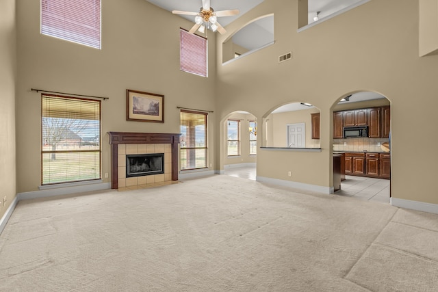 unfurnished living room with light carpet, visible vents, arched walkways, a tile fireplace, and ceiling fan