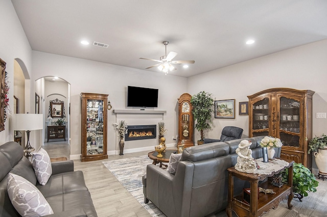 living room with arched walkways, ceiling fan, visible vents, a lit fireplace, and light wood finished floors