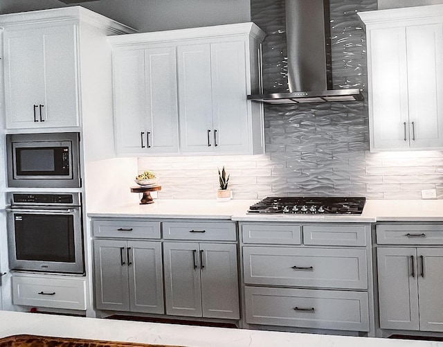 kitchen with gray cabinets, stainless steel appliances, light countertops, wall chimney exhaust hood, and tasteful backsplash