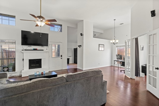 living room with baseboards, dark wood-style flooring, a brick fireplace, high vaulted ceiling, and ceiling fan with notable chandelier