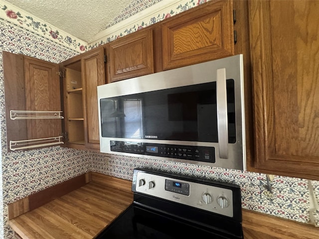 kitchen with a textured ceiling, stainless steel appliances, wood finished floors, brown cabinetry, and wallpapered walls