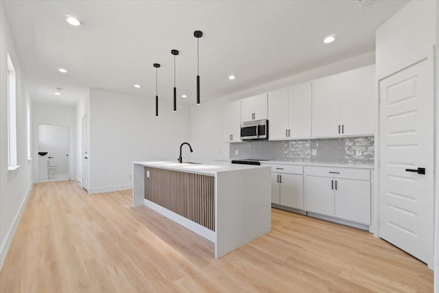 kitchen with tasteful backsplash, stainless steel microwave, a kitchen island with sink, light countertops, and light wood-type flooring