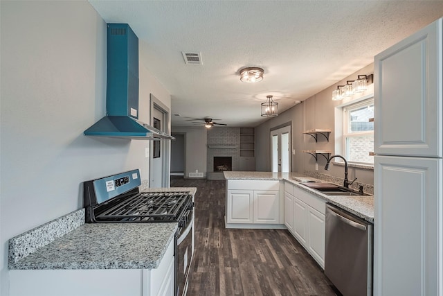 kitchen featuring a sink, visible vents, wall chimney range hood, appliances with stainless steel finishes, and a brick fireplace