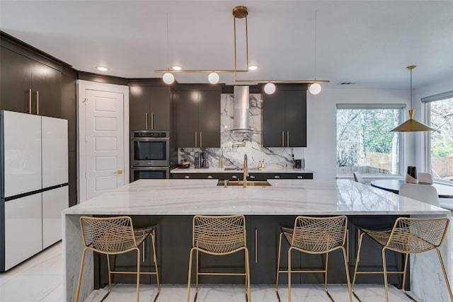 kitchen featuring decorative backsplash, freestanding refrigerator, stainless steel double oven, wall chimney range hood, and a sink