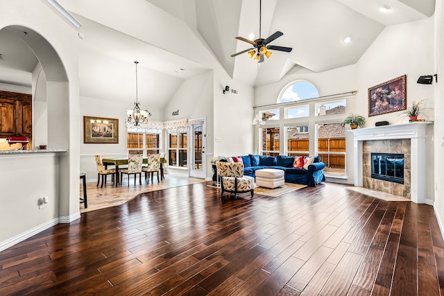 living room with baseboards, a tiled fireplace, wood finished floors, high vaulted ceiling, and ceiling fan with notable chandelier