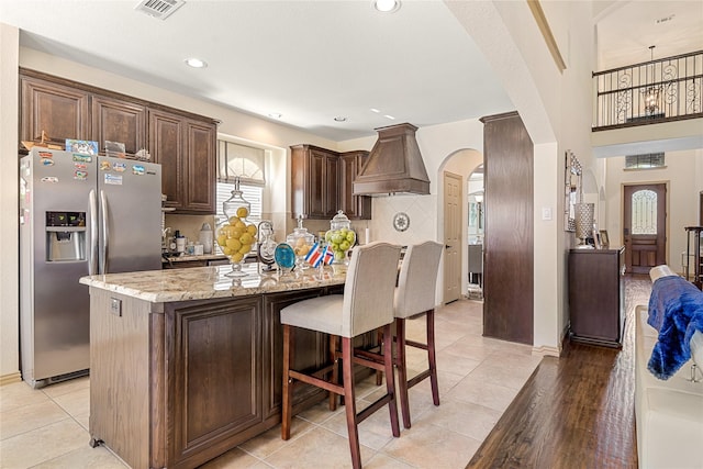 kitchen featuring arched walkways, stainless steel fridge with ice dispenser, custom range hood, light stone counters, and dark brown cabinets