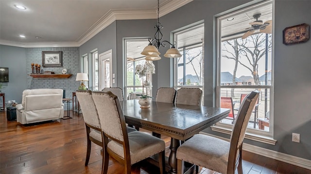 dining room featuring crown molding, wood-type flooring, a brick fireplace, baseboards, and ceiling fan with notable chandelier