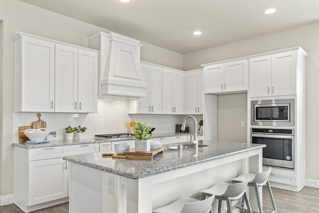 kitchen featuring decorative backsplash, appliances with stainless steel finishes, white cabinets, a sink, and premium range hood