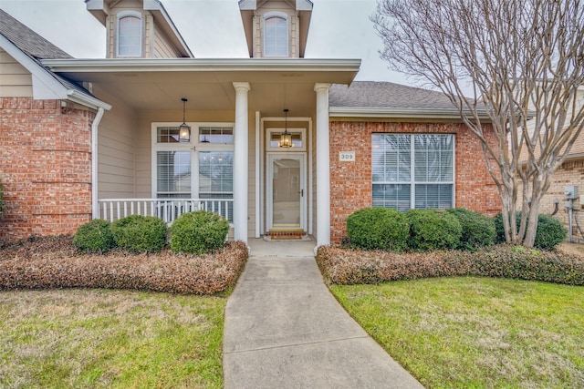 doorway to property with covered porch, brick siding, a lawn, and roof with shingles