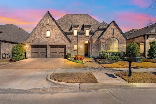 french country home with a garage, concrete driveway, brick siding, and roof with shingles