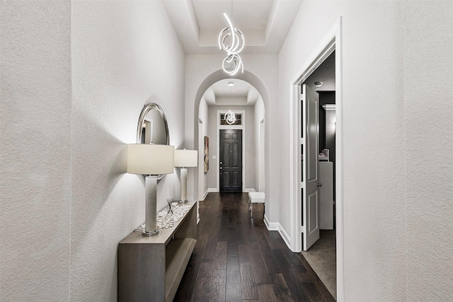 hallway featuring a tray ceiling, dark wood-style flooring, arched walkways, a textured wall, and baseboards