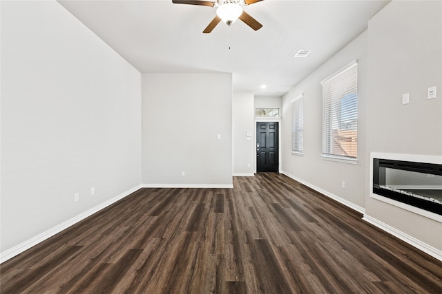 unfurnished living room featuring dark wood-type flooring, a glass covered fireplace, ceiling fan, and baseboards