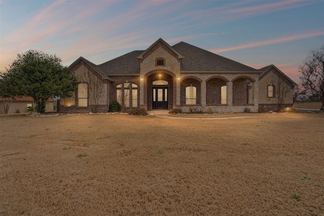 french country inspired facade with a shingled roof, brick siding, and a yard