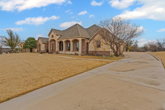 french country home featuring stone siding, a shingled roof, concrete driveway, and a front yard