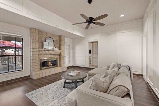 living room featuring a ceiling fan, a textured ceiling, dark wood finished floors, a fireplace, and a decorative wall