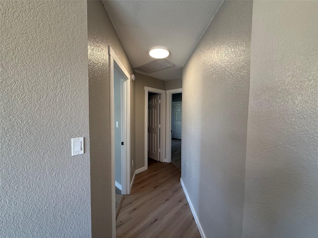 hall featuring a textured ceiling, a textured wall, wood finished floors, visible vents, and baseboards