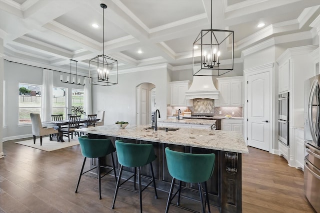 kitchen featuring arched walkways, custom exhaust hood, decorative backsplash, a sink, and an island with sink