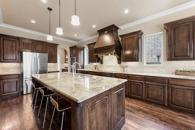 kitchen featuring stainless steel fridge, a center island with sink, arched walkways, dark wood-style flooring, and premium range hood