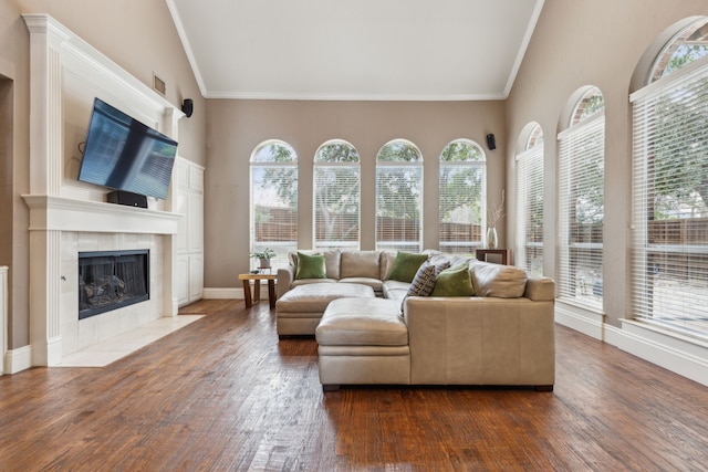 living area with dark wood-style flooring, a tiled fireplace, ornamental molding, high vaulted ceiling, and baseboards