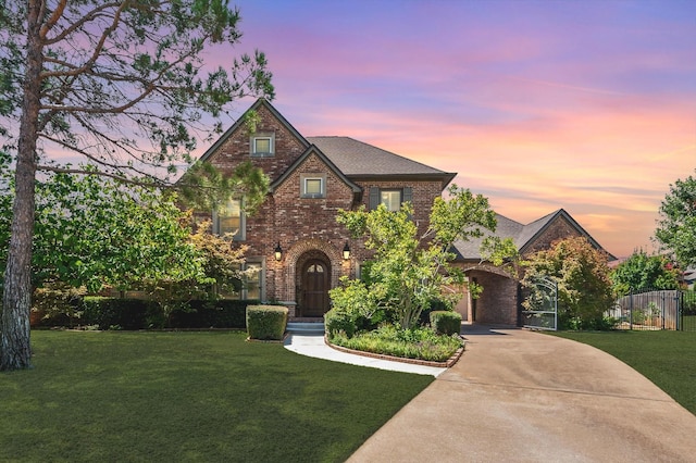 tudor home featuring brick siding, a shingled roof, concrete driveway, fence, and a front lawn