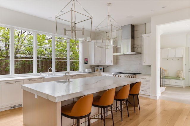 kitchen featuring a kitchen island with sink, a sink, light wood-type flooring, decorative backsplash, and wall chimney exhaust hood