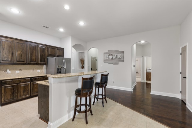 kitchen with arched walkways, a kitchen island with sink, stainless steel refrigerator with ice dispenser, decorative backsplash, and a kitchen bar