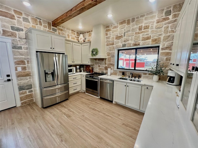 kitchen with light wood finished floors, custom range hood, beamed ceiling, stainless steel appliances, and a sink