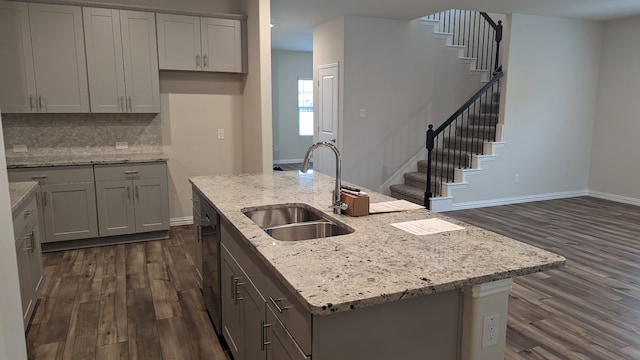 kitchen featuring a kitchen island with sink, gray cabinetry, a sink, backsplash, and dark wood finished floors