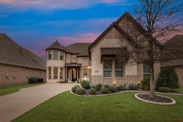 french country home with a shingled roof, concrete driveway, stone siding, a yard, and brick siding