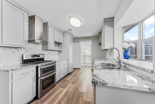 kitchen featuring a sink, light wood-style floors, wall chimney range hood, double oven range, and tasteful backsplash